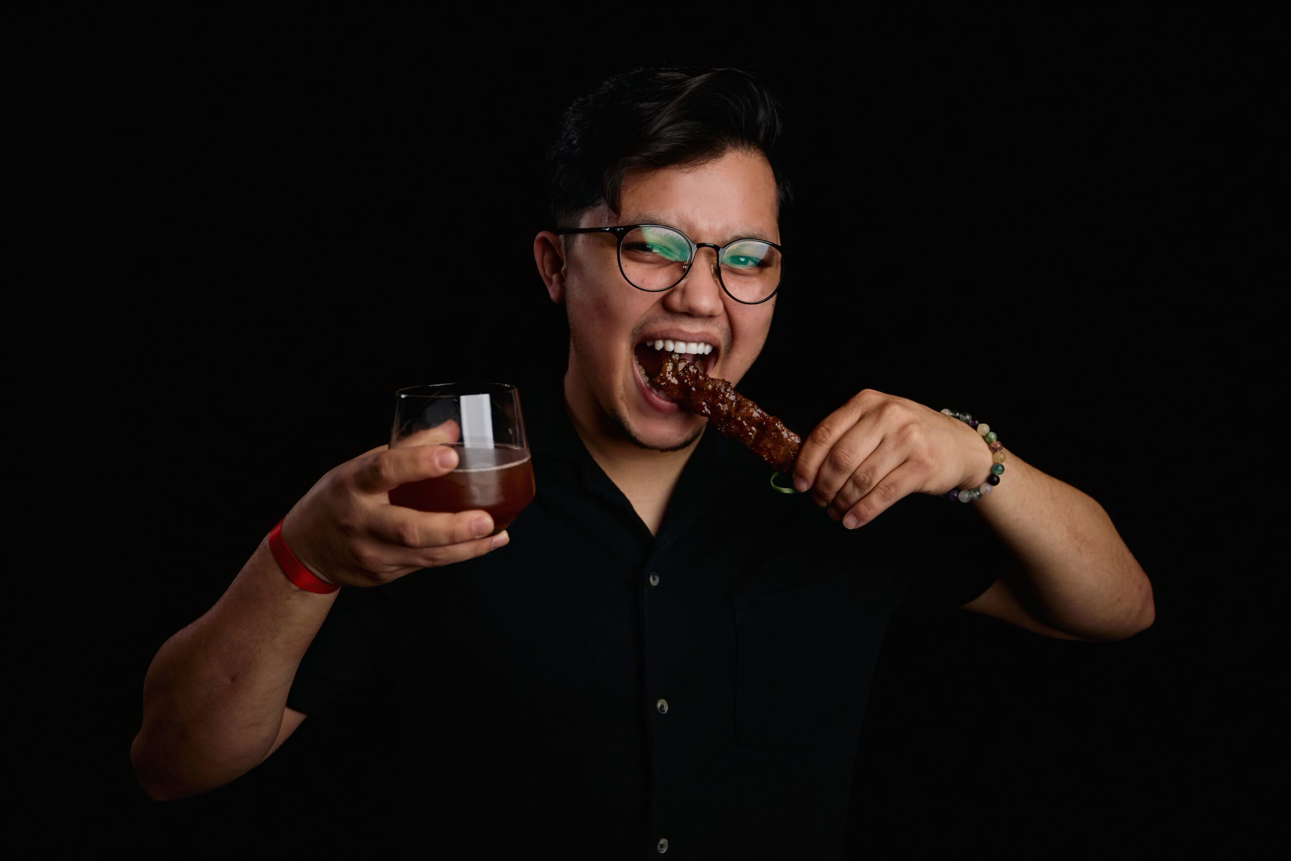 Person with short dark hair and glasses, smiling, holding a glass of drink and a large piece of meat, against a black background.