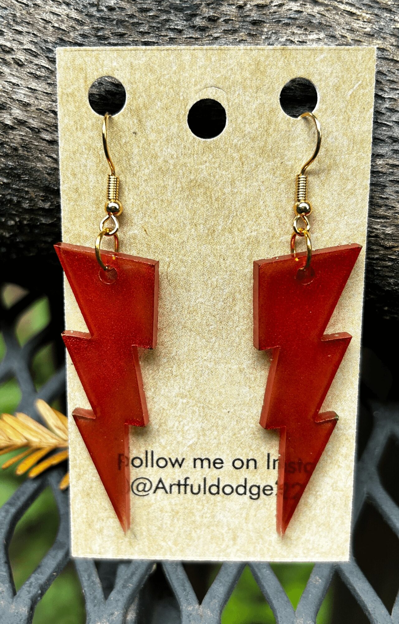 Red lightning bolt earrings on a card with text, against a dark background with plants visible.