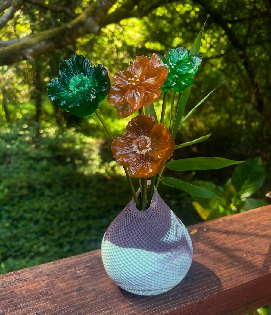 Glass flower bouquet in a white vase on a wooden surface outdoors with green trees in background.
