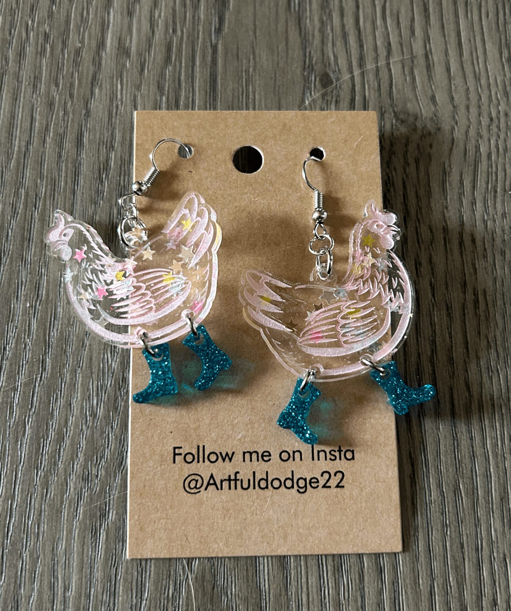 Clear chicken-shaped earrings with blue feet on a brown card, on a wooden surface. Text: Follow me on Insta @Artfuldodge22.