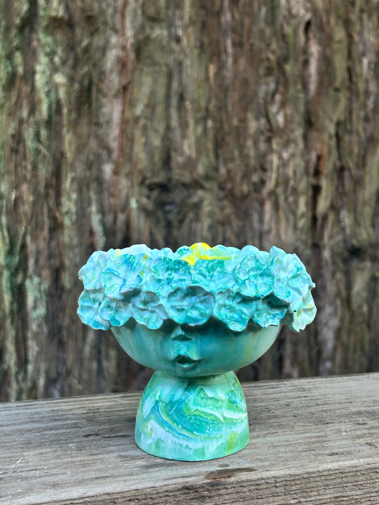 Decorative ceramic head with blue flowers as hair, on a wooden surface, with a textured background.