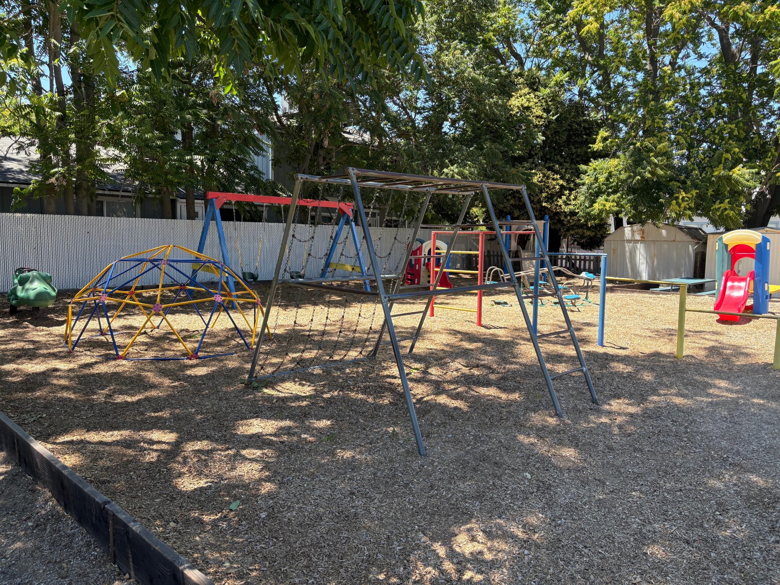 Playground with swings, slide, and climbing structures under trees, with shadows on the ground.