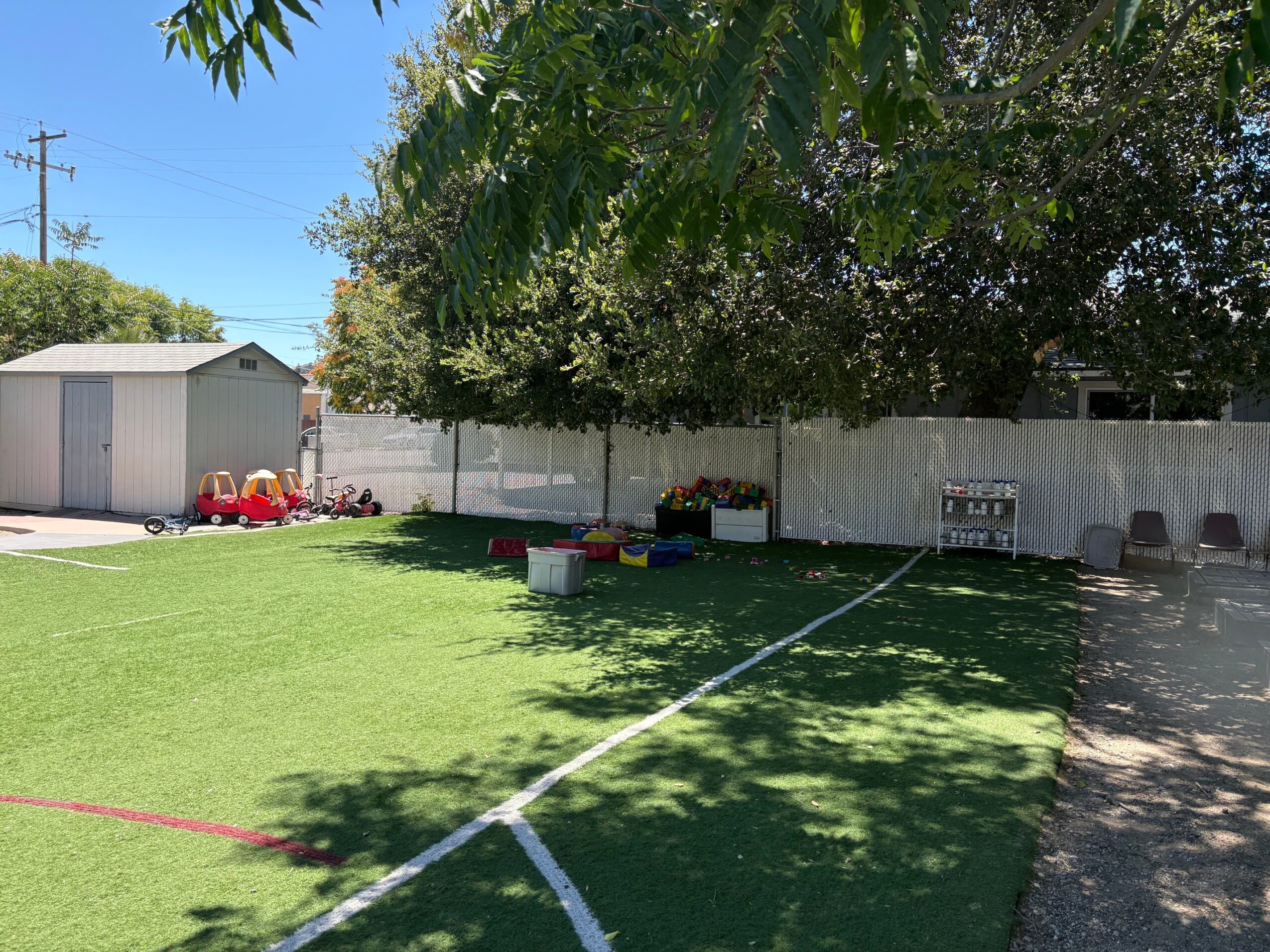 Backyard with green grass, trees, a white fence, and a shed, with toys and a bench visible.
