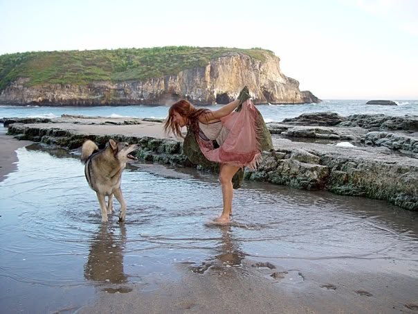 Girl in pink dress playing with dog on beach near rocks and cliff, water and sky in background.