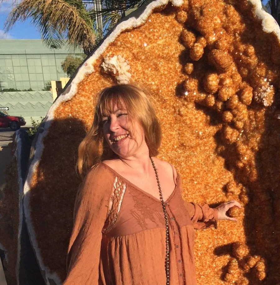 Woman smiling outdoors, standing next to a large, textured, orange-brown object, with trees and a building in the background.