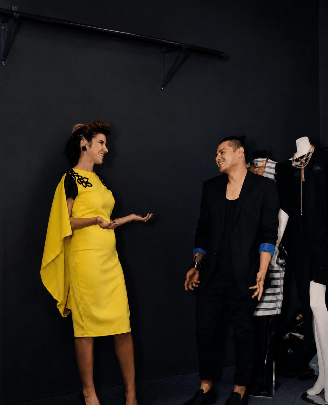 Two women are talking in a dark room, one in a yellow dress and the other in black, with clothing on a rack nearby.