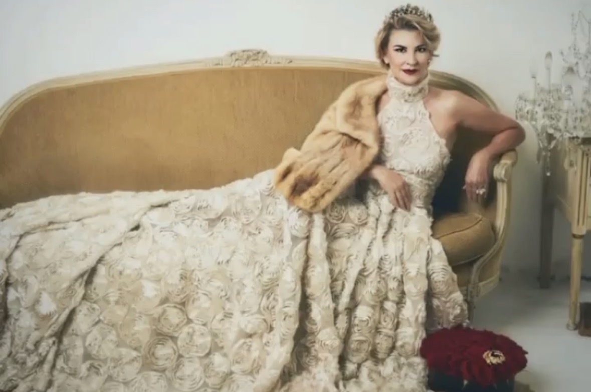 Woman in vintage dress and fur stole reclining on a yellow sofa in a decorated room.