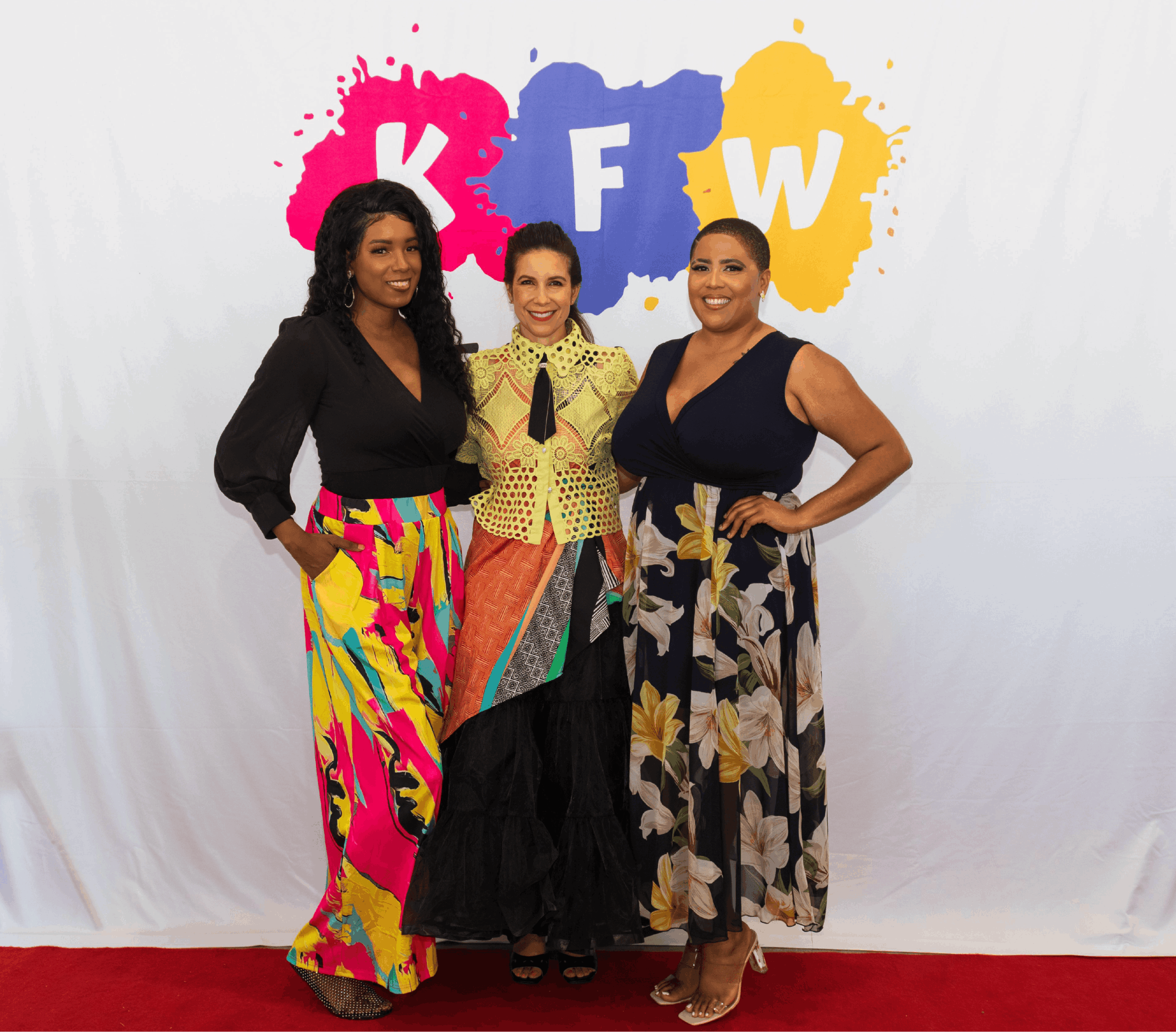 Three women standing in front of a white backdrop with colorful KFW letters, smiling and posing for the photo.