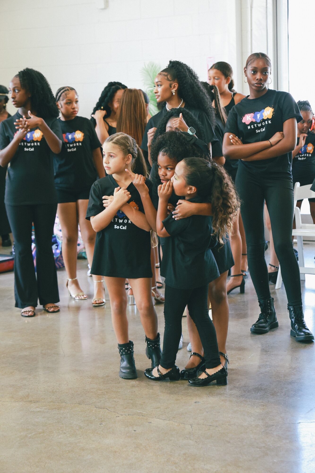 Group of young girls standing indoors, some with arms crossed, others with hands clasped, wearing matching black shirts.