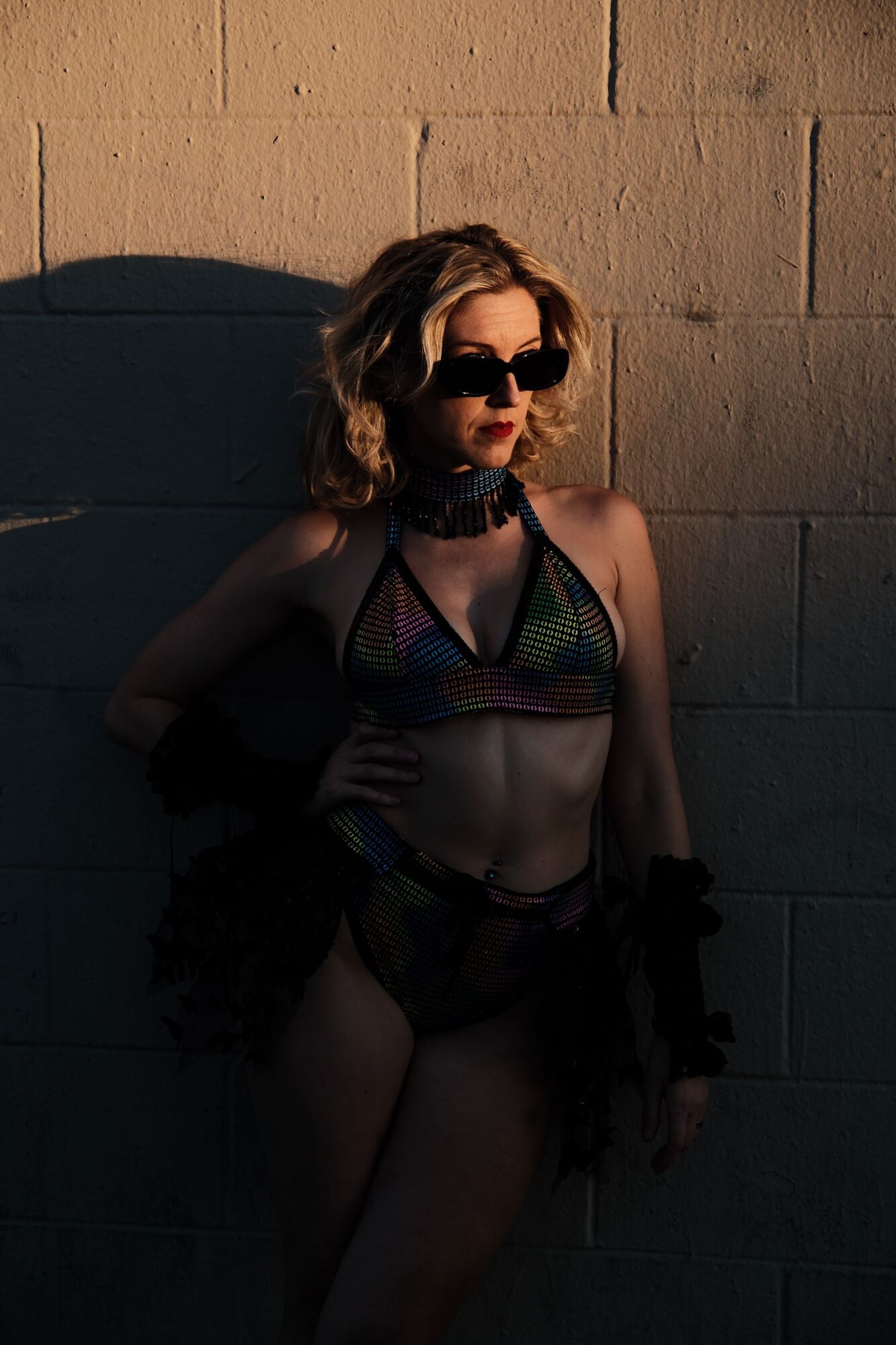 Woman in sunglasses and colorful swimsuit leaning against a wall, sunlight casting shadows, outdoor setting.