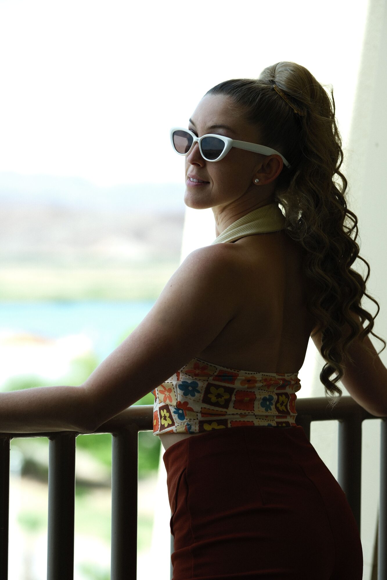 Woman lying sideways on a bed, wearing sunglasses and a colorful top, with a blurred window background.