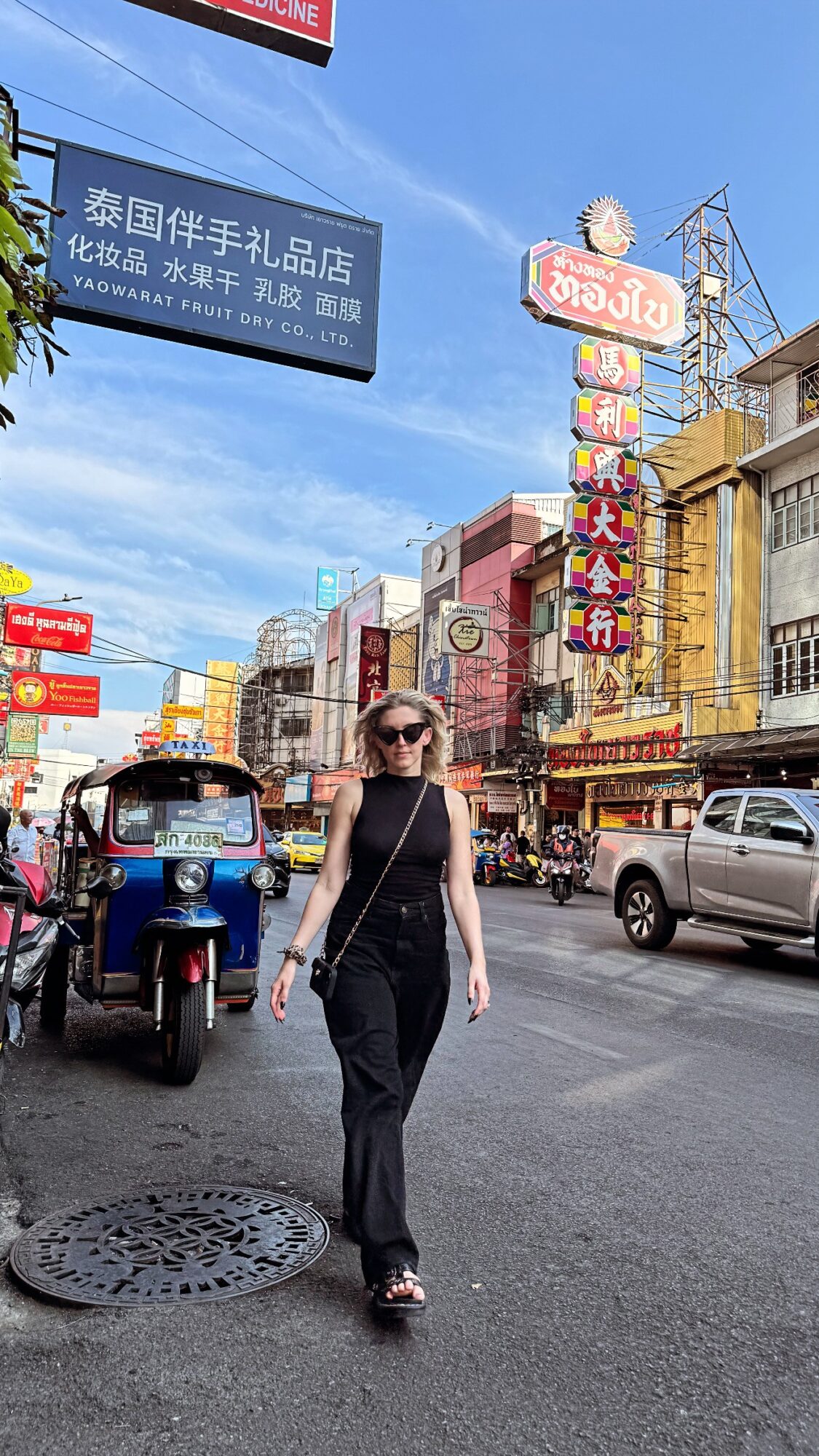 Woman walking on busy street with shops, signs, and vehicles under blue sky.