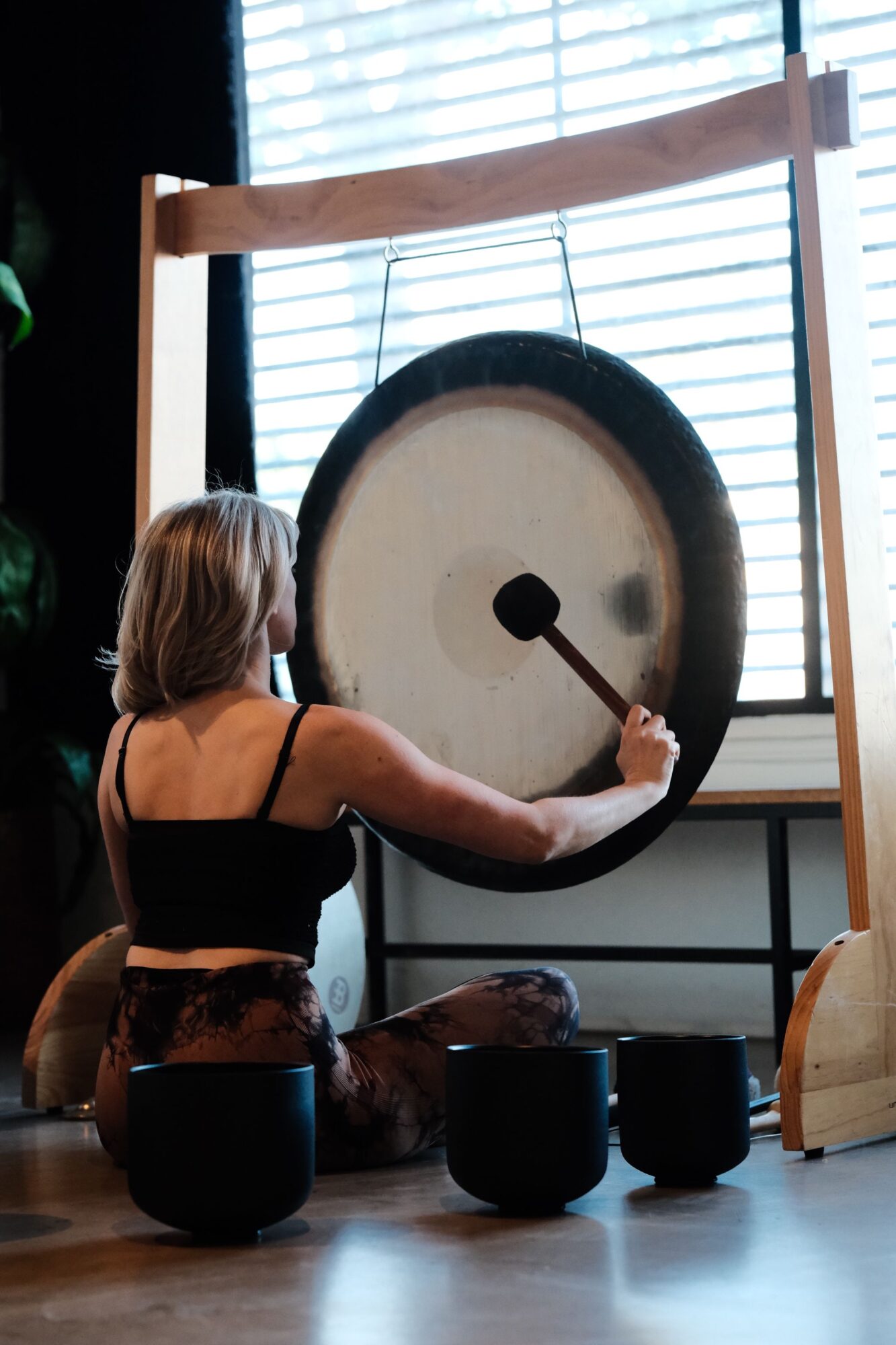 Person hitting a gong with a mallet in a room with large windows and wooden floors.