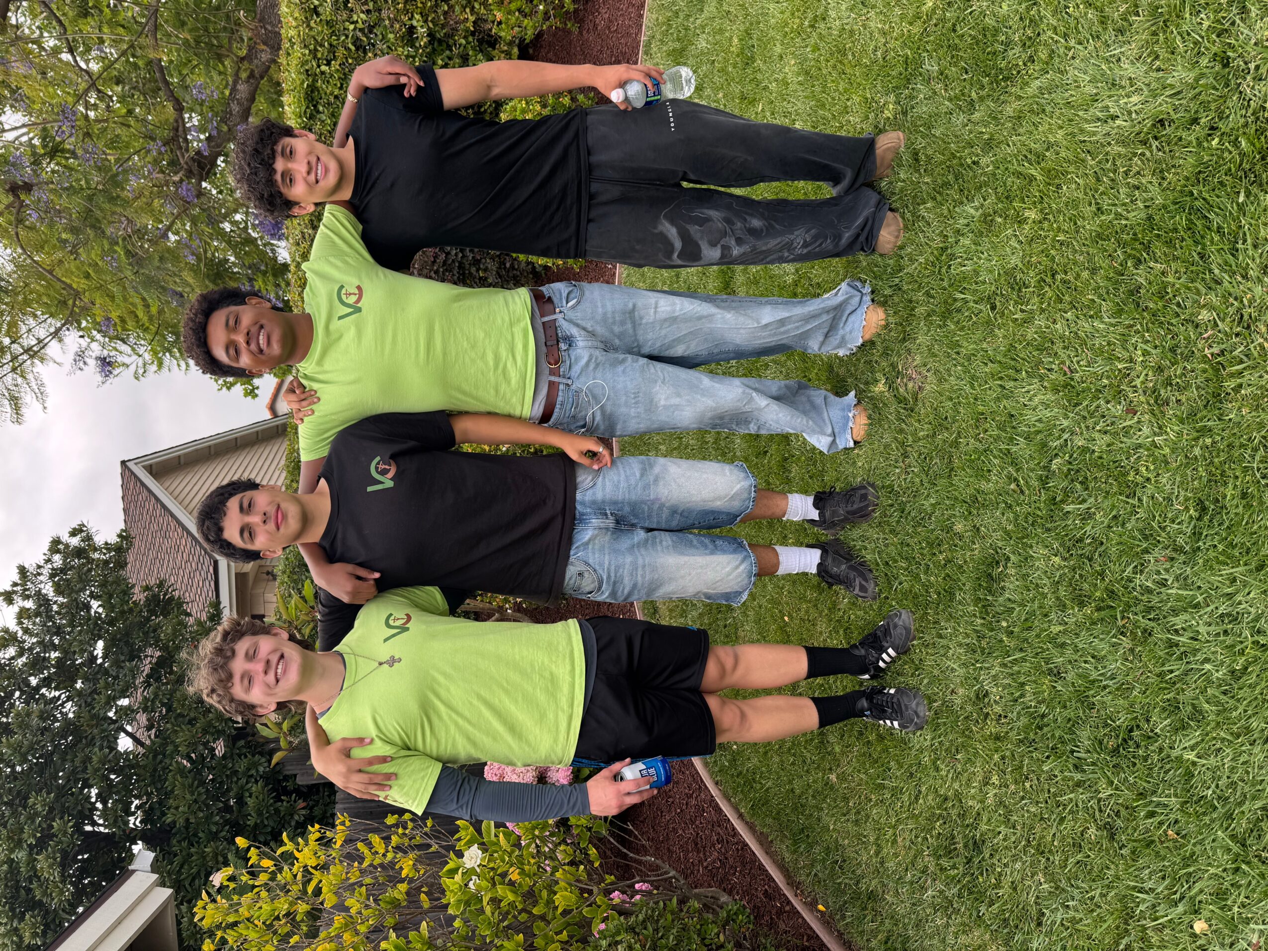 Four people standing on grass in front of a house, two wearing neon green shirts, two in black shirts, with trees and a house in the background.