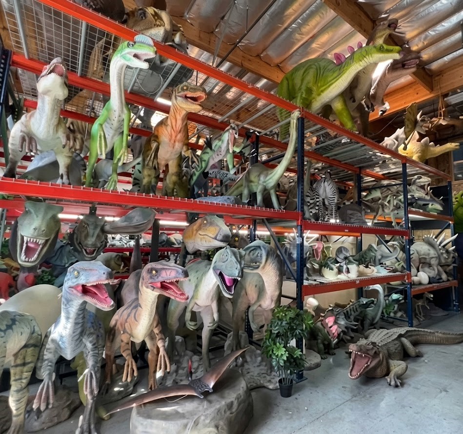 Shelves filled with various dinosaur models in a storage area, with some plants and a wooden ceiling visible.