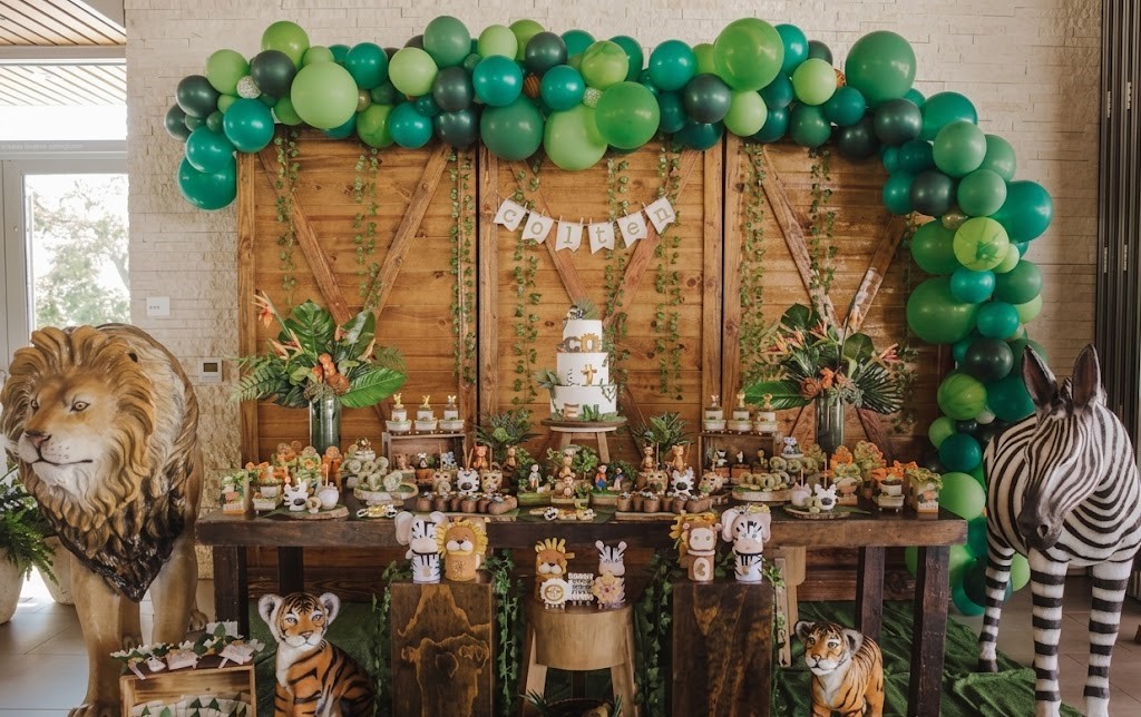 Party table with a cake, animal decorations, and green balloon arch, featuring a lion and zebra in the scene.