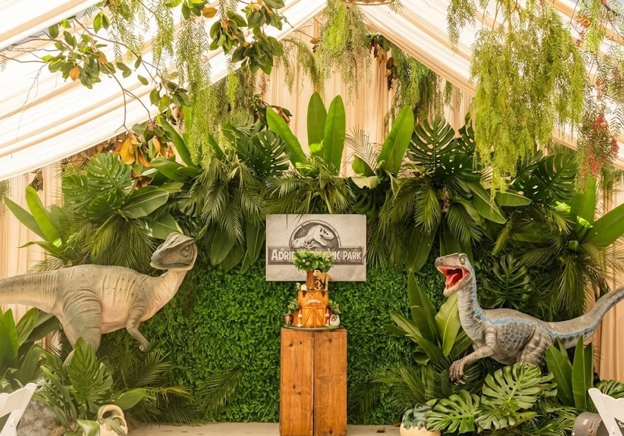 Dinosaur models displayed among green plants and a sign, with a wooden pedestal in front, inside a greenhouse or exhibit space.