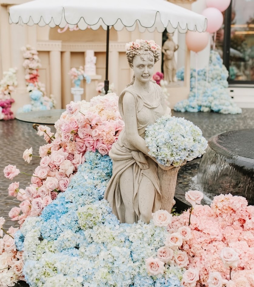 Statue of a woman holding a bouquet, surrounded by pink and blue flowers, with an umbrella overhead, outdoors.