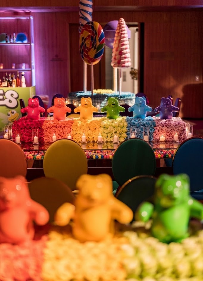 Colorful gummy bears arranged on a table with multicolored chairs, a wooden wall, and party decorations in the background.