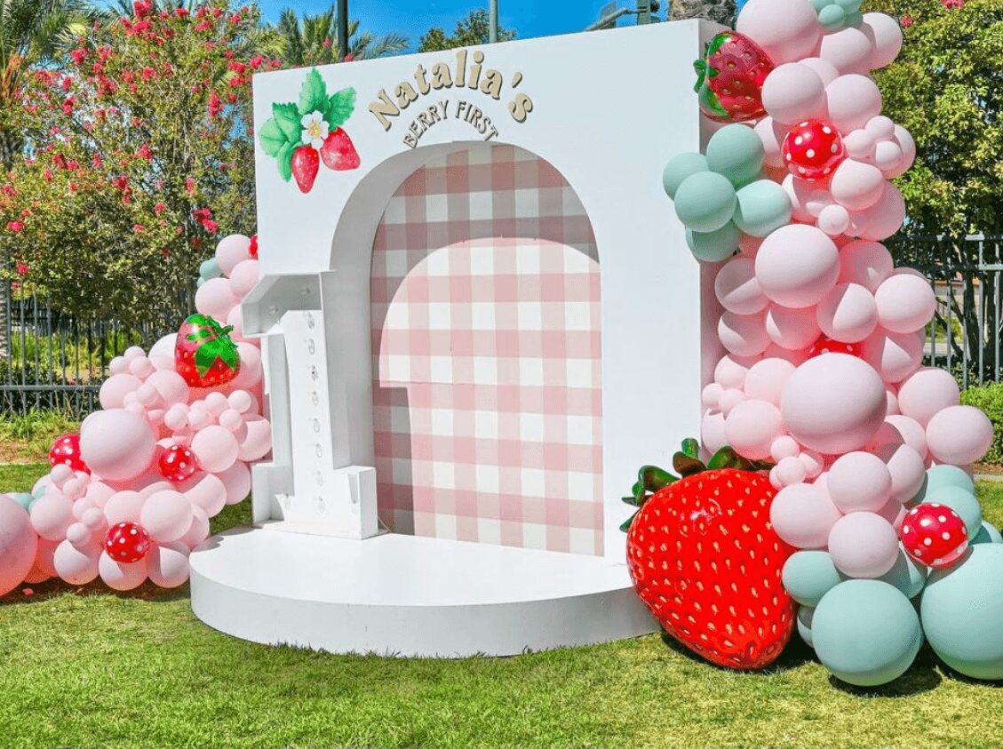 Decorative backdrop with pink, white, and green balloons, a large strawberry, and a checkered arch for a celebration.