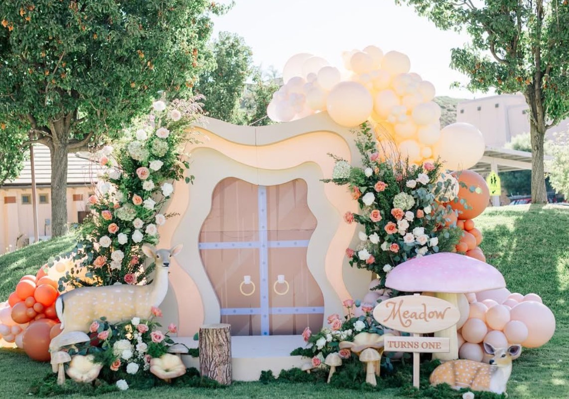 Decorative arch with flowers and balloons, set outdoors with trees and grass, for celebration or event.