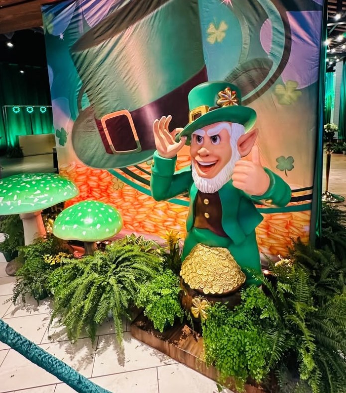 Leprechaun character with a green hat and suit, giving a thumbs-up, surrounded by plants and a colorful backdrop.