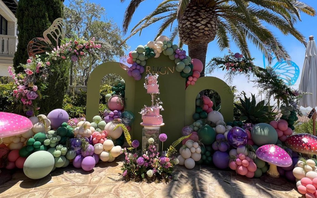 Decorative setup with colorful balloons, flowers, and a central cake, outdoors with palm trees and blue sky.