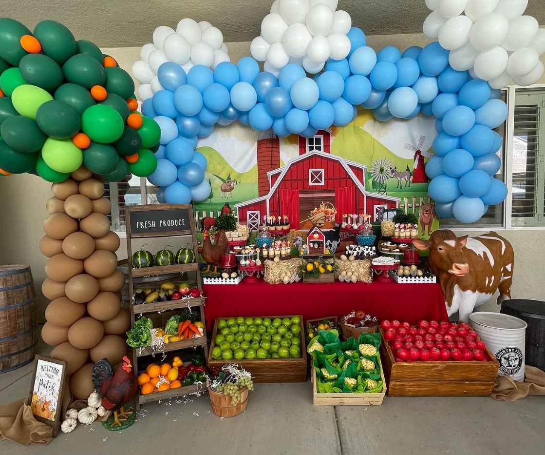 Decorative farm-themed display with balloon arch, tree, and table of farm produce and treats, set outdoors with a barn backdrop.