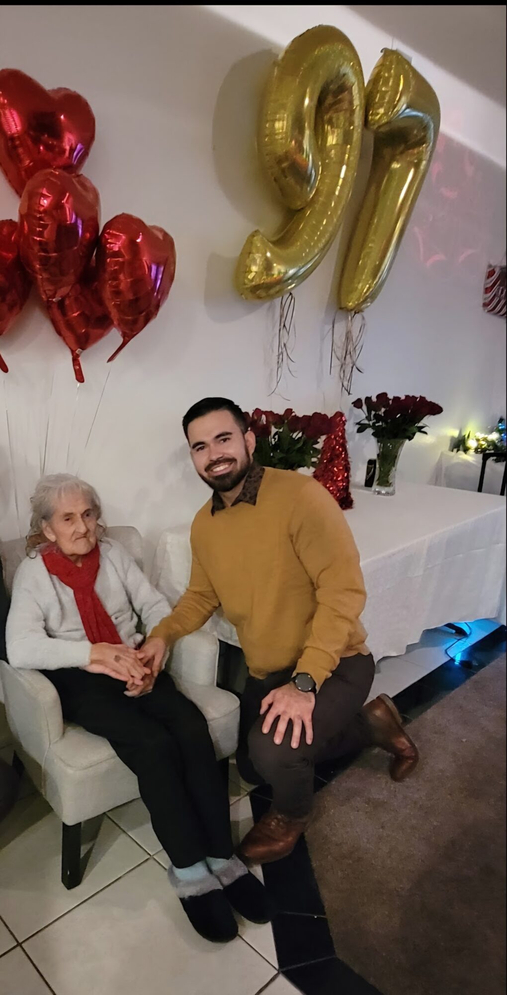 A young man and an elderly woman sit together, holding hands, with balloons and decorations in the background.