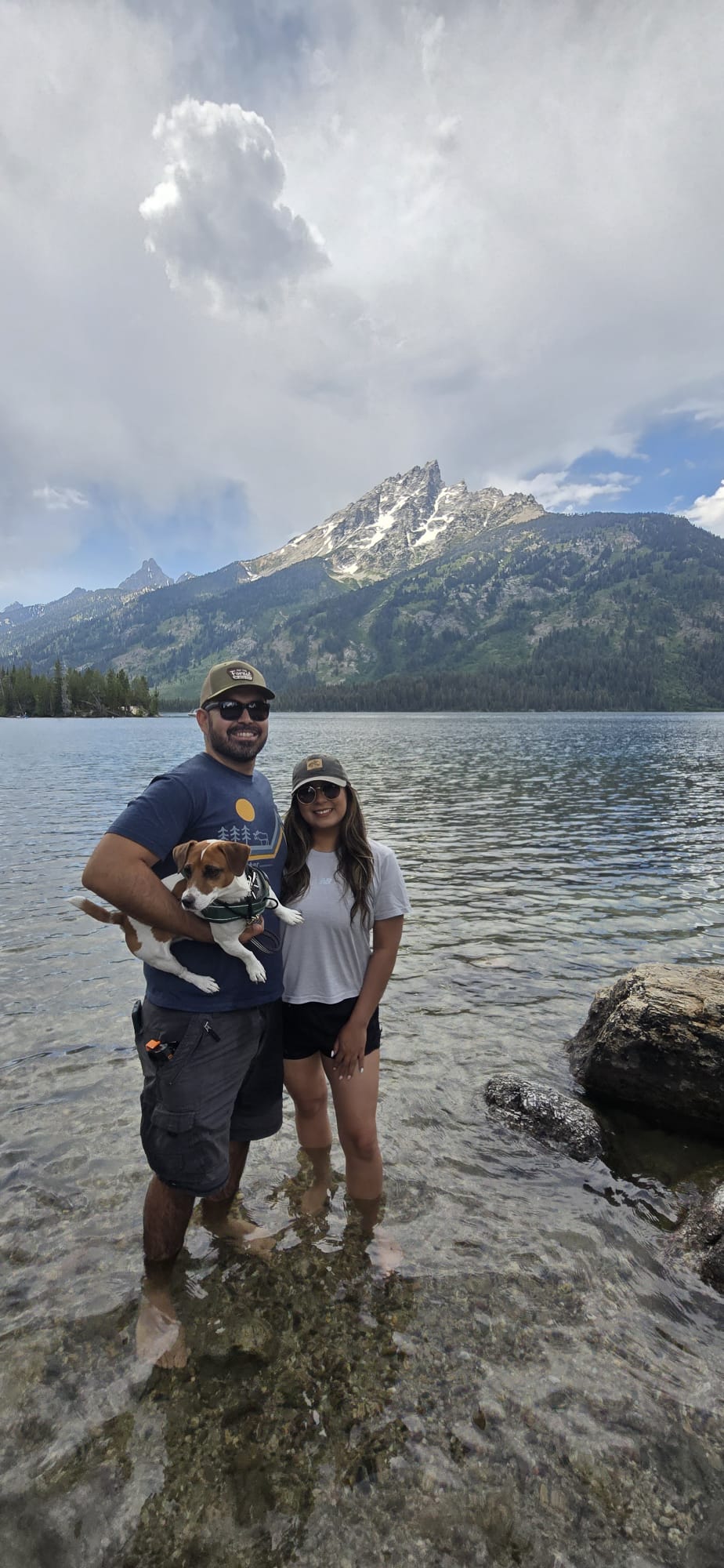 Two people stand in a lake with mountains and cloudy sky in the background, holding a dog between them.
