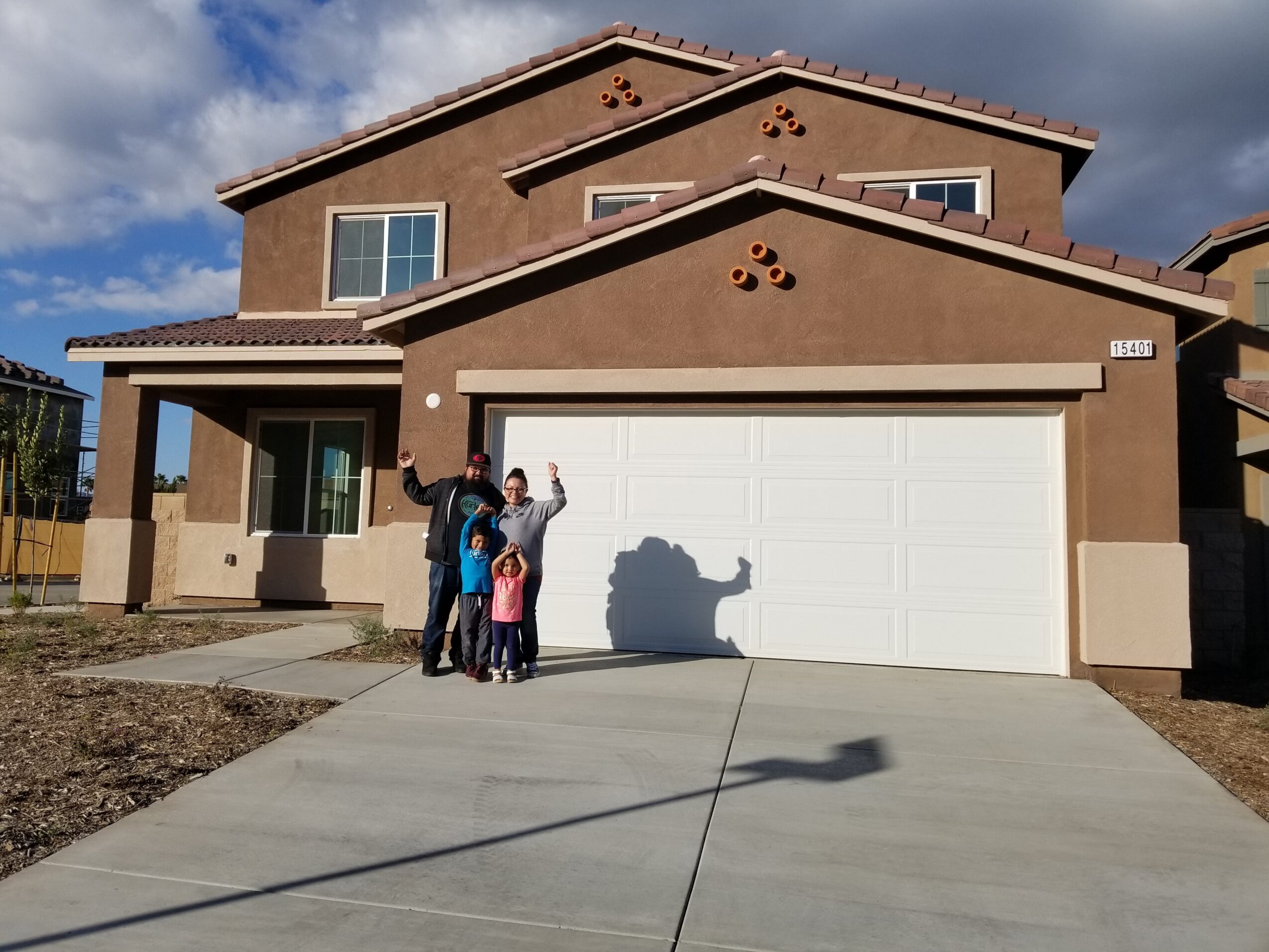 Group of four people standing in front of a two-story house with a white garage door, under a partly cloudy sky.