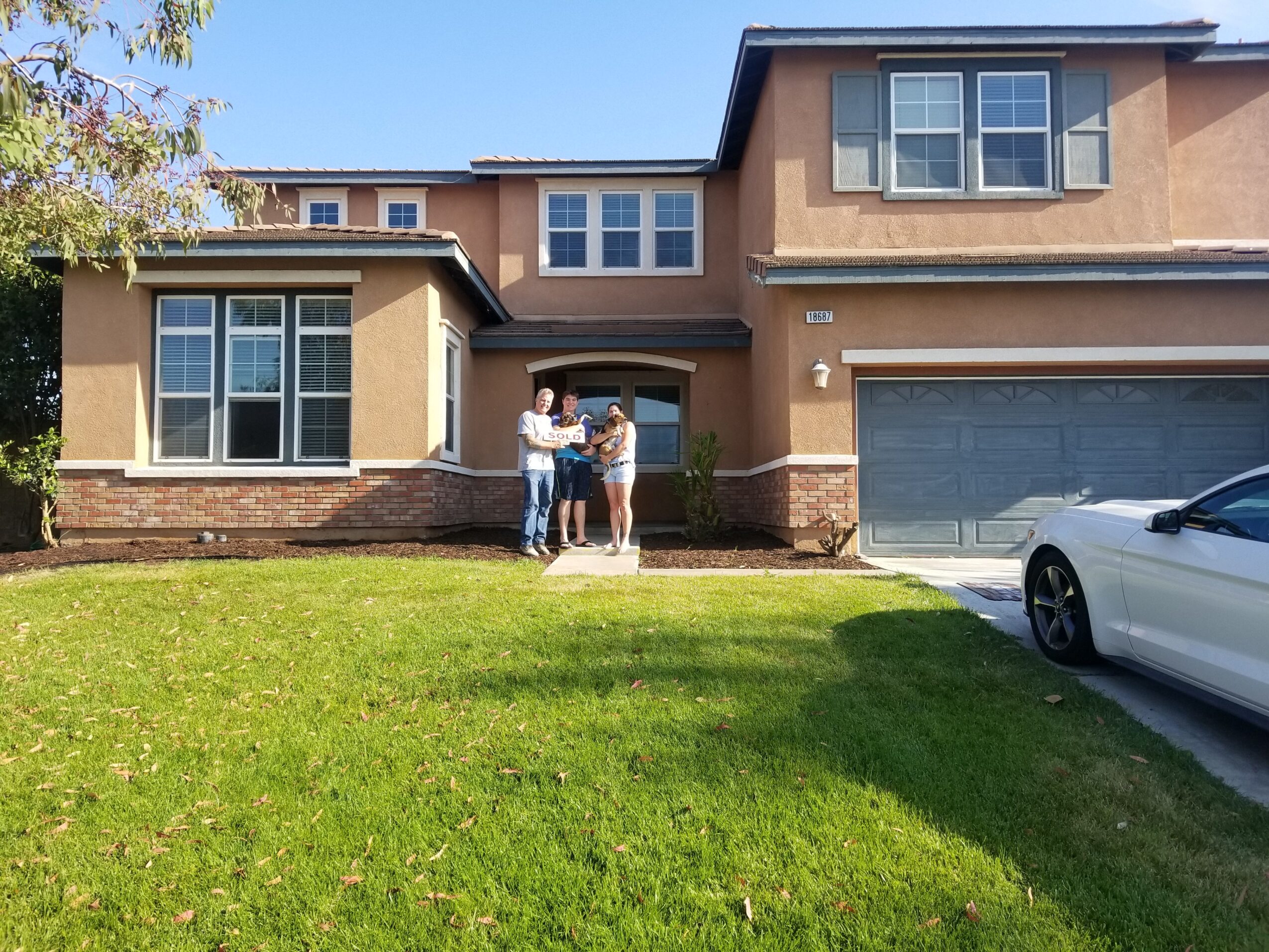 Two people stand in front of a two-story house with a garage, on a grassy lawn under a clear sky.
