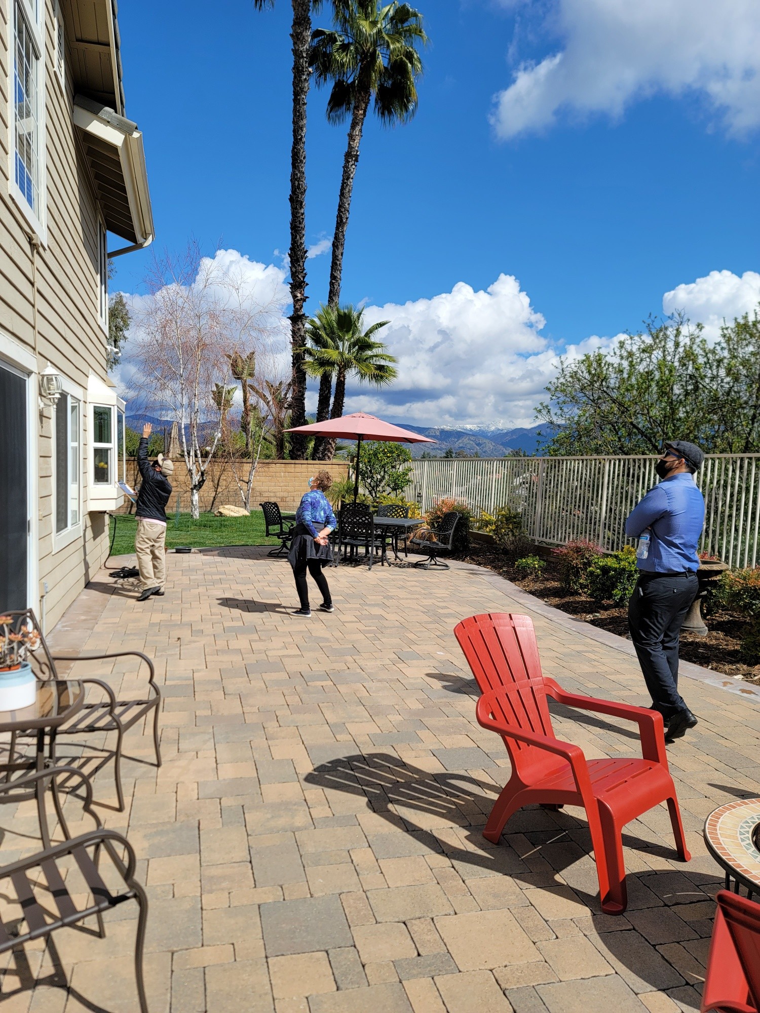 People in a backyard with trees, a fence, and outdoor furniture under a blue sky with clouds.