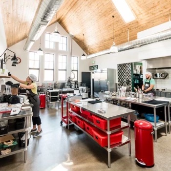 Two people working in a spacious, well-lit commercial kitchen with high wooden ceiling, large windows, and stainless steel equipment.