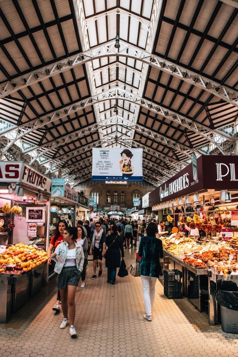 Indoor market with high arched ceiling, people shopping at fruit stalls, signs for various stores, and a digital screen in the center.