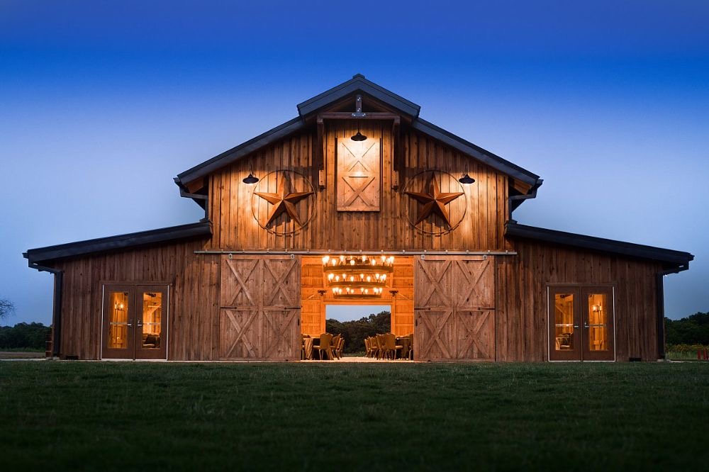 Large wooden barn with open central doorway, illuminated interior, and small windows, set against a twilight sky with a grassy foreground.