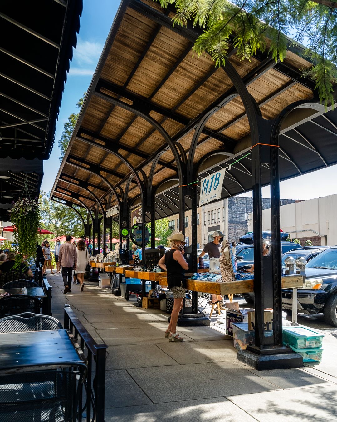 Outdoor market with wooden canopy, people shopping at tables, parked cars, and trees in the background.