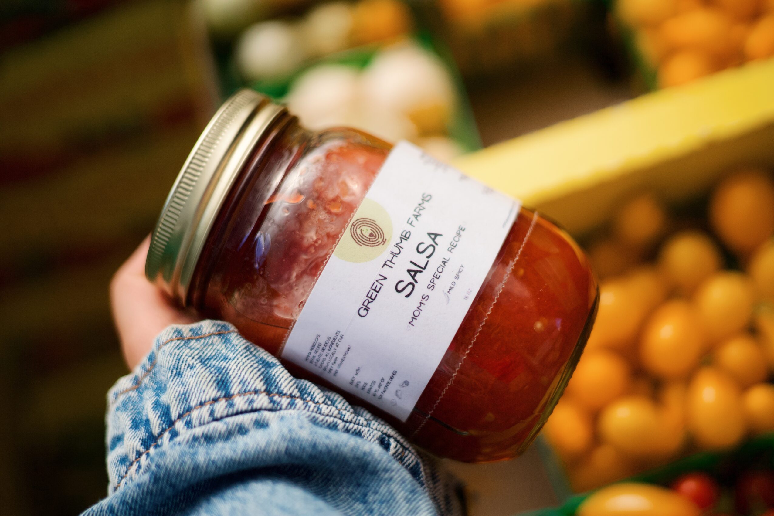 Person holding a jar of red salsa in a grocery store, with produce in the background.