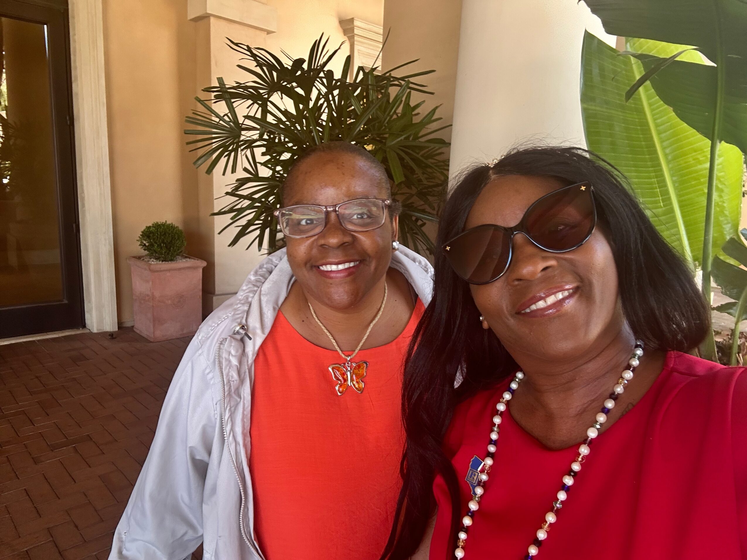 Two women smiling, one wearing glasses and a red top, the other with sunglasses and a red dress, outdoors with plants.