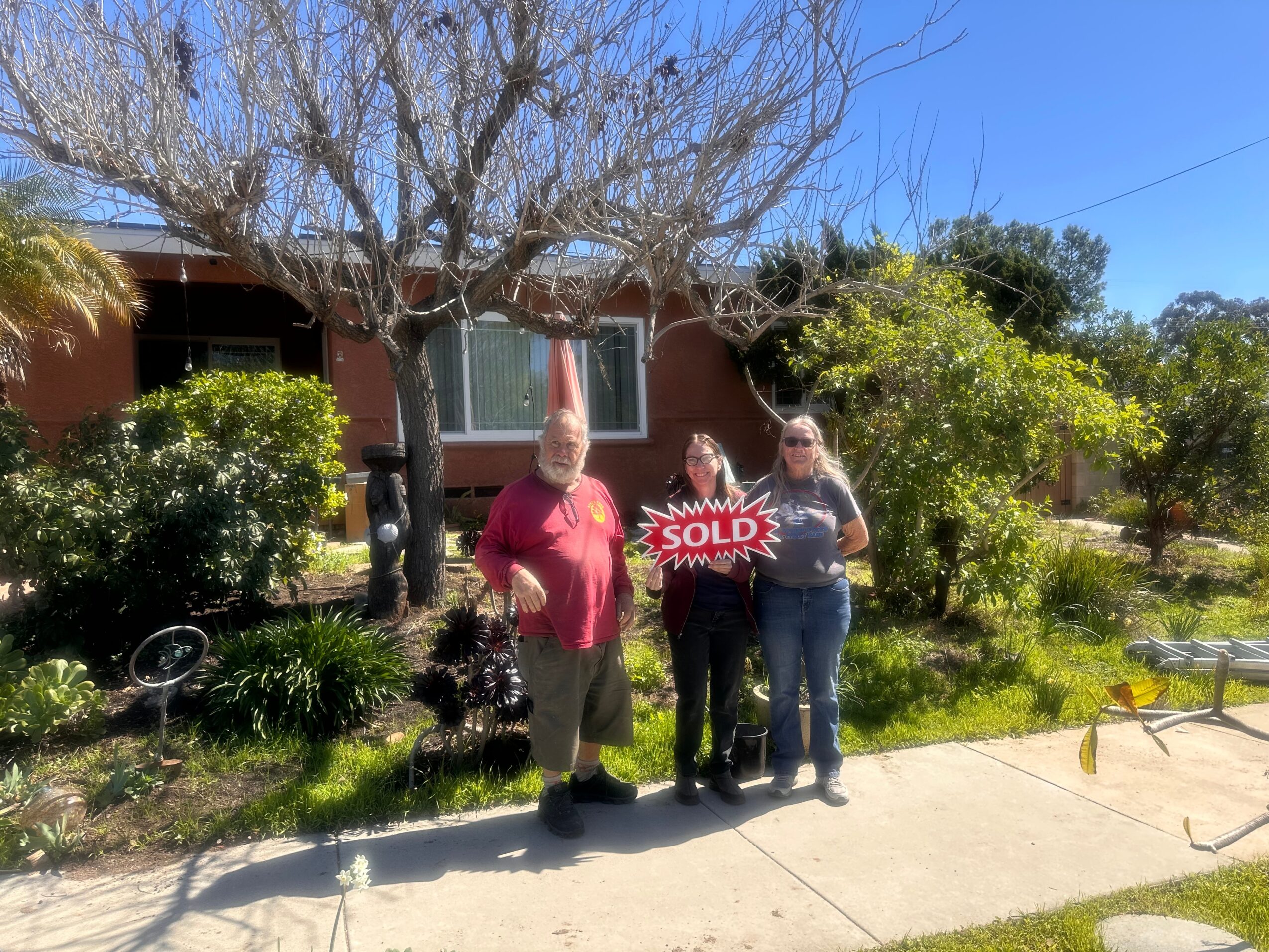 Three people standing outside a house with a tree and garden, one holding a 'SOLD' sign, on a sunny day.