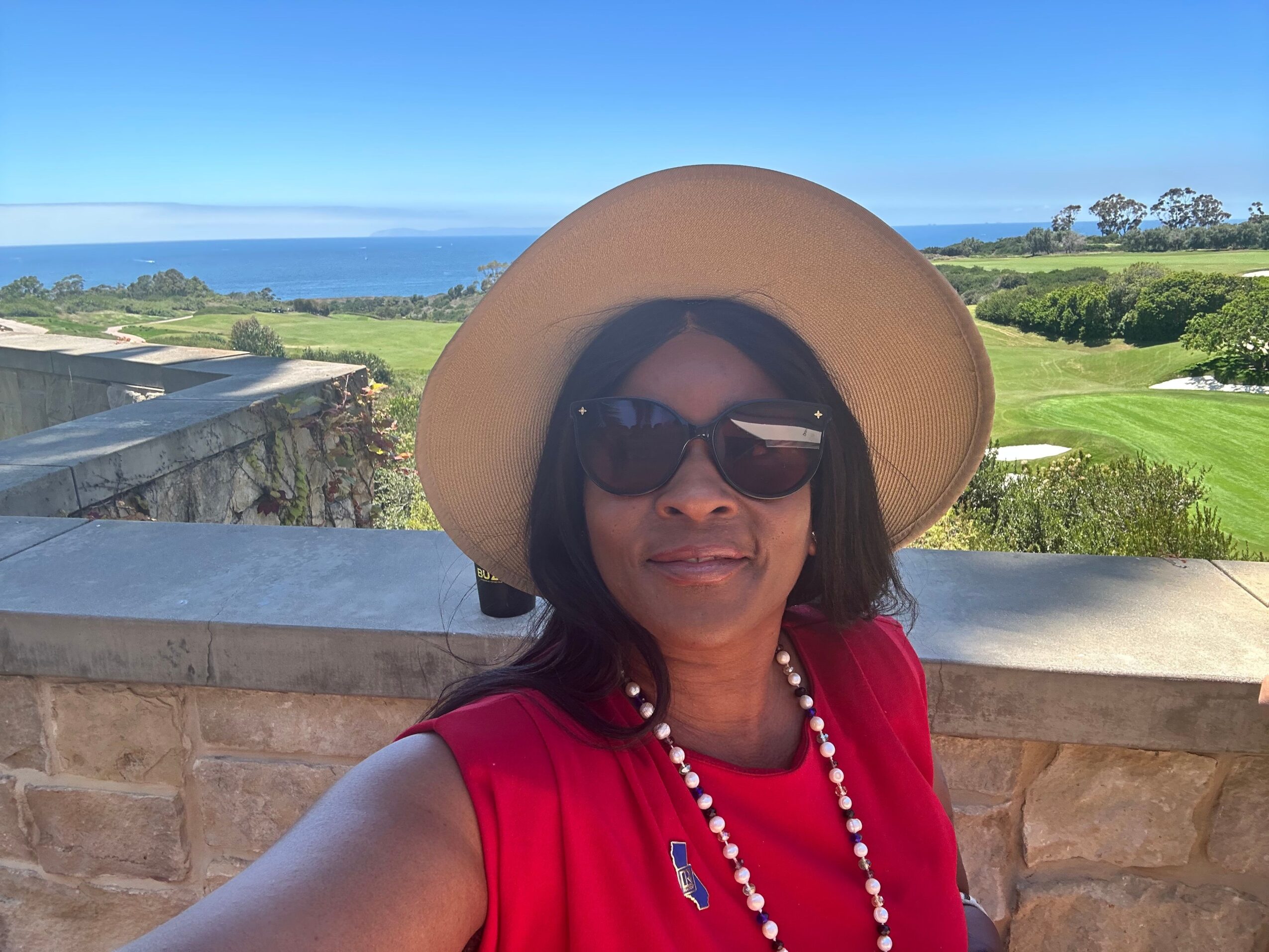 Woman wearing a wide-brimmed hat and sunglasses, smiling outdoors with a blue sky and green landscape in the background.