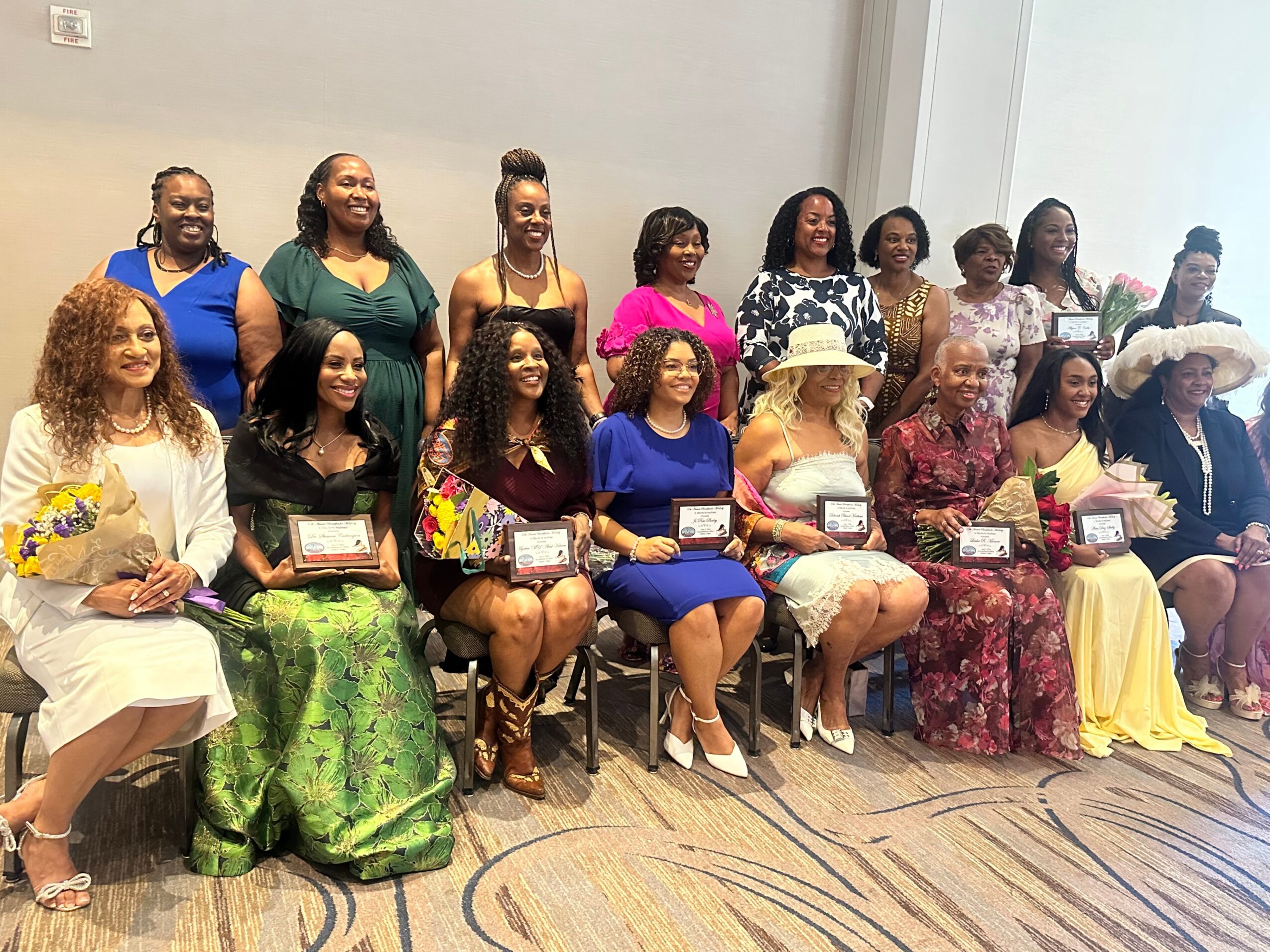 Group of women in colorful dresses, some holding awards or certificates, posing indoors against a plain wall.