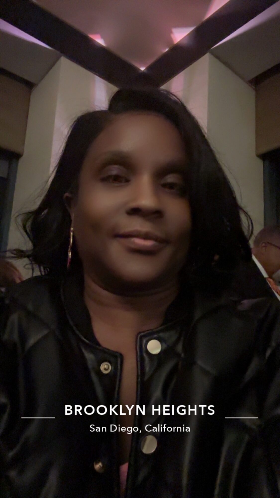 Woman with dark hair and earrings taking a selfie indoors, with a ceiling light and people in the background.