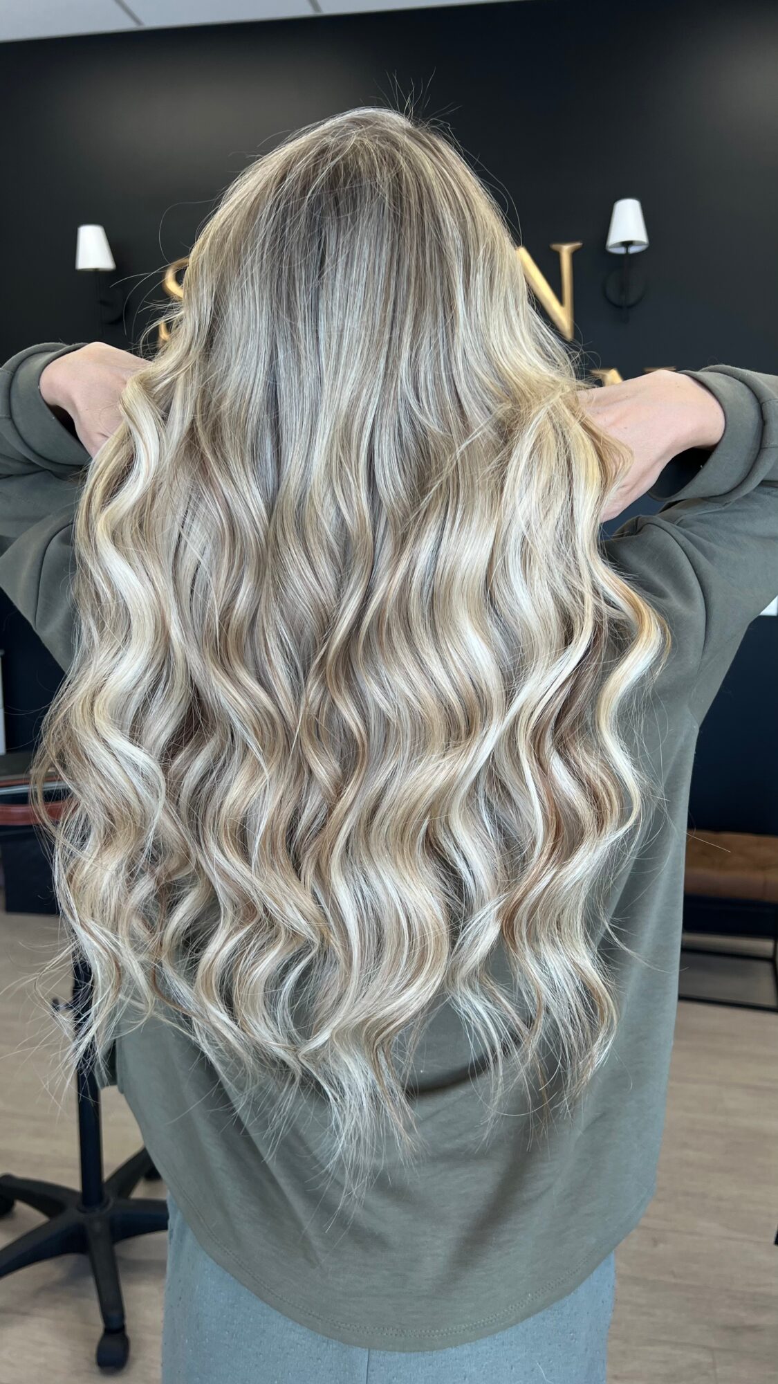 Back view of a person with long, wavy, blonde hair, wearing a gray top, in an indoor setting.