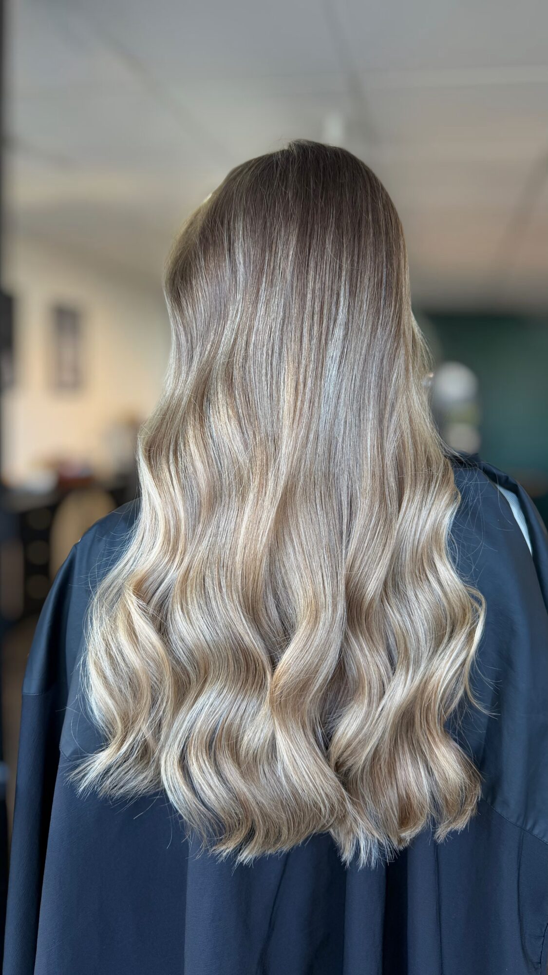 Back view of a person with long, wavy, blonde hair, wearing a dark top or gown.