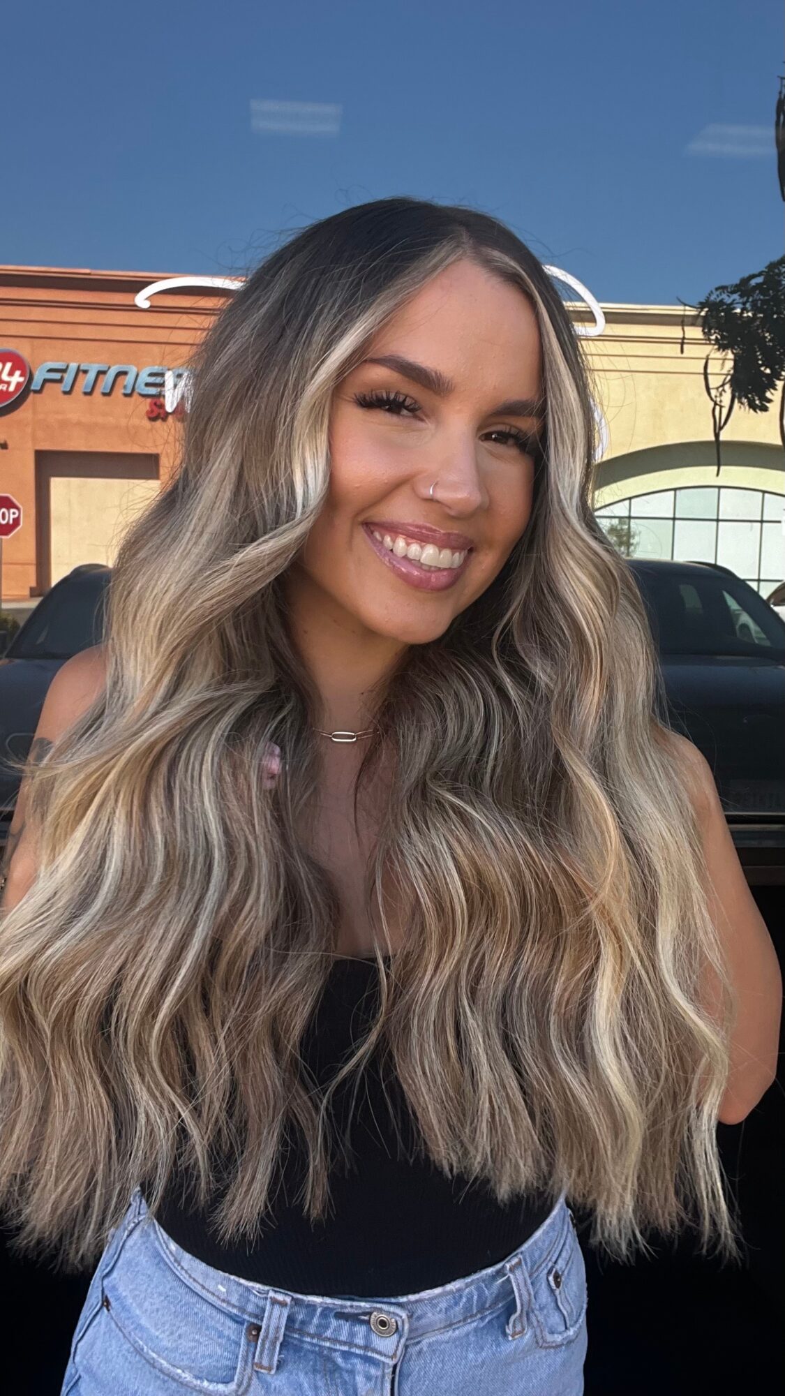 Smiling woman with long wavy hair standing outdoors in front of a building and parked cars.