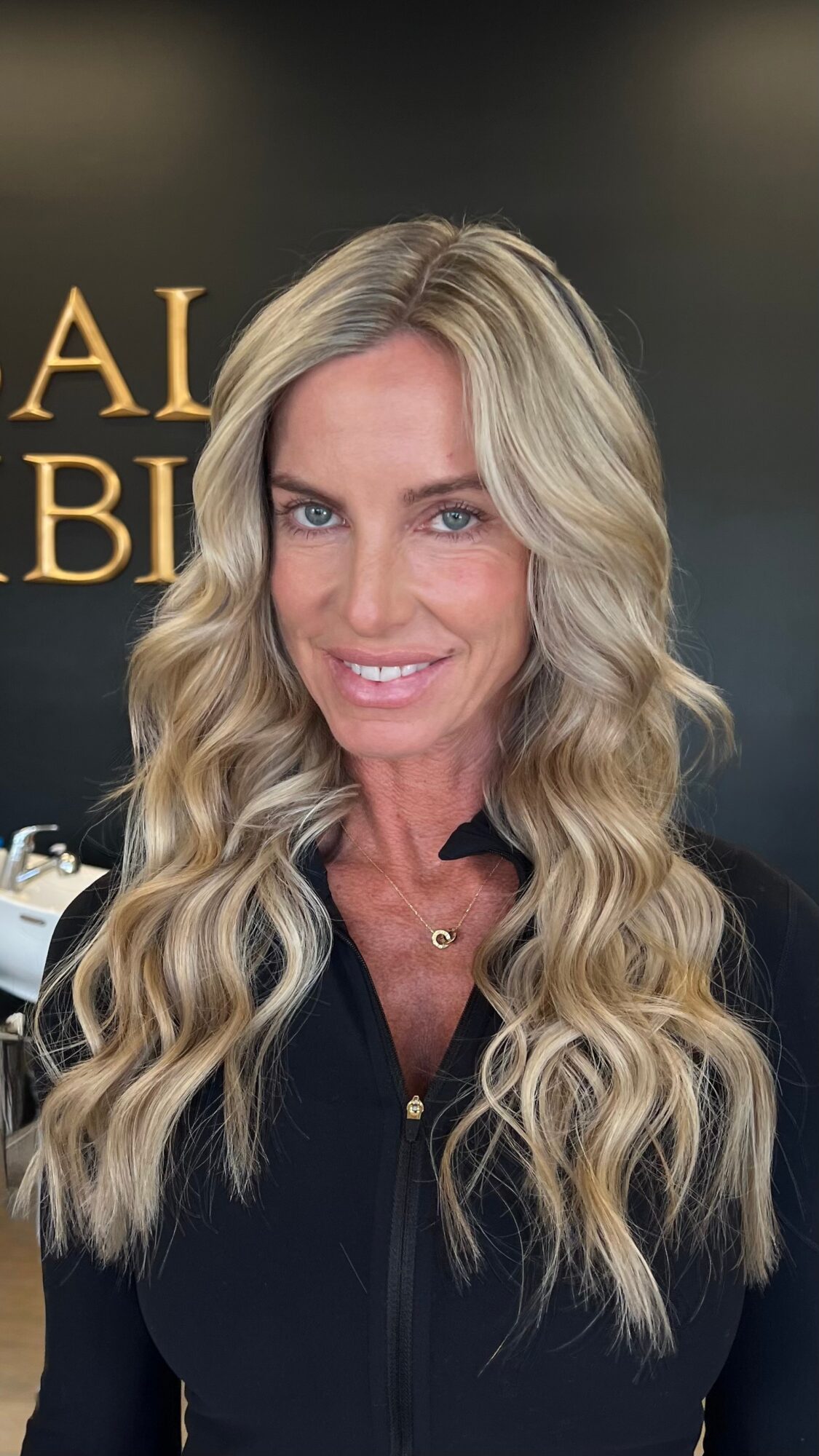 Woman with long wavy blonde hair smiling, wearing a black top, in front of a dark background with gold text