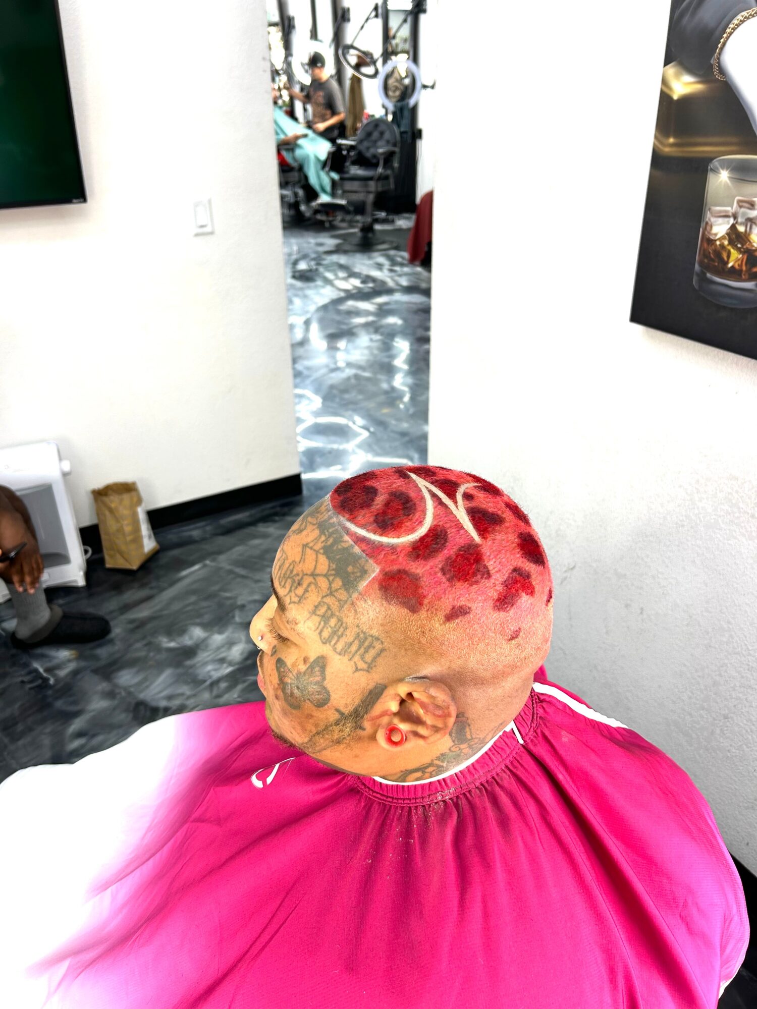 Person with a shaved head featuring tattoos and red paint, wearing a pink cape, in a room with white walls and artwork.