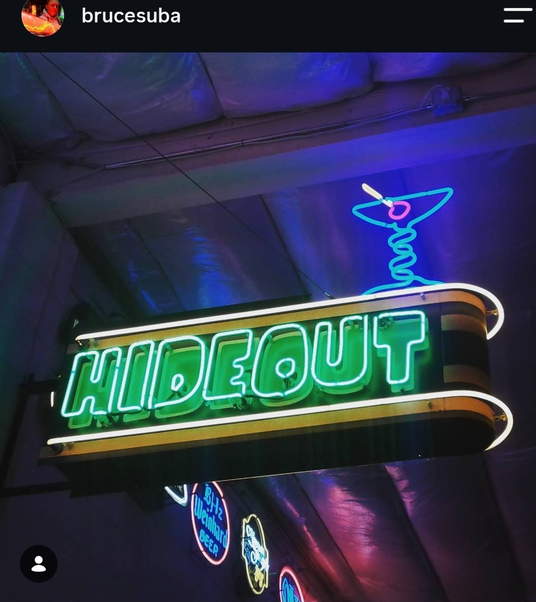 Neon sign reading 'HIDEOUT' with a cocktail glass illustration above, illuminated in green, blue, and pink colors.