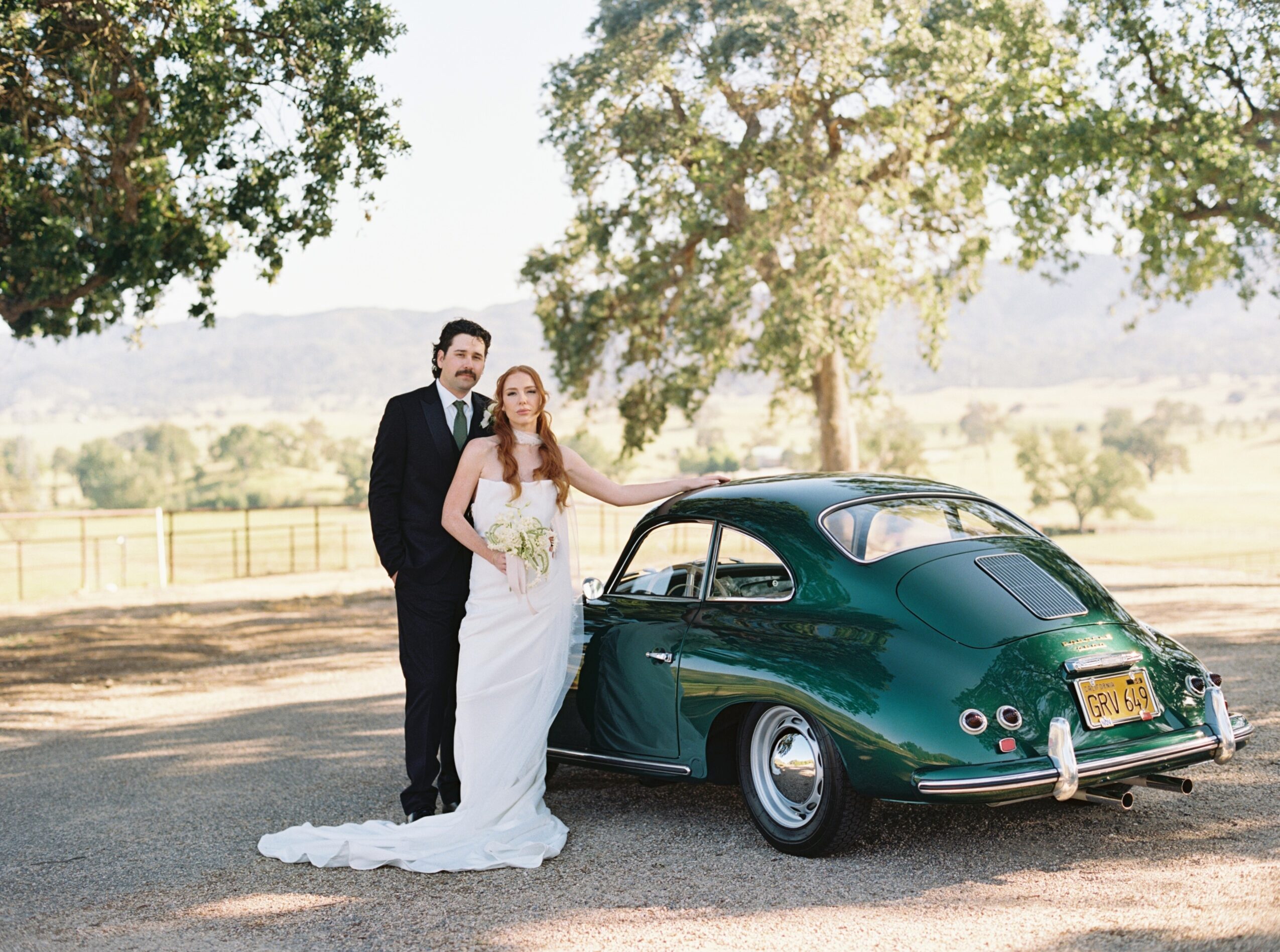 Bride and groom stand beside a vintage green car outdoors with trees and a landscape in the background.